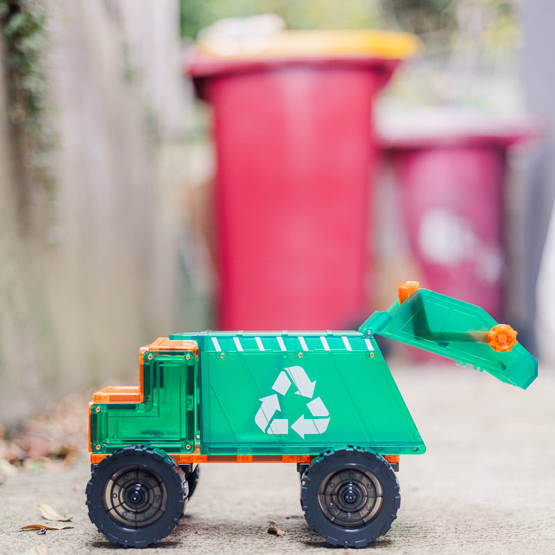 Toy magnetic tile  garbage truck with recycling symbol in front of real trash bins.
